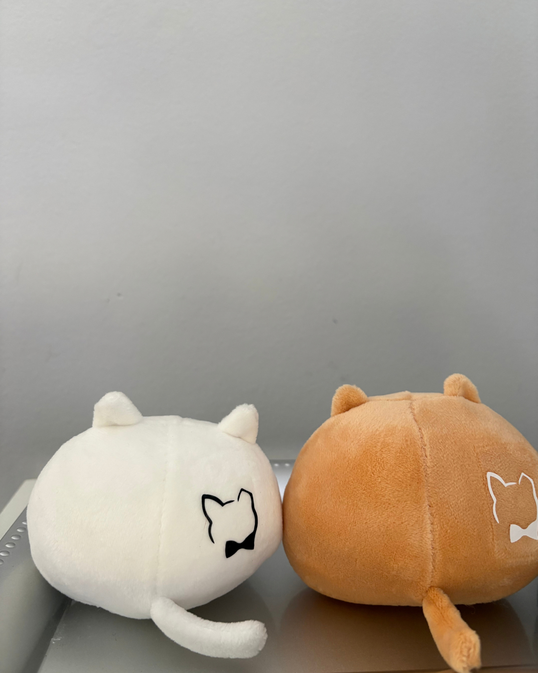 Two round plush toys photographed on a shelf against a plain gray wall. The left plush is white with a black cat face outline and bow-tie detail.