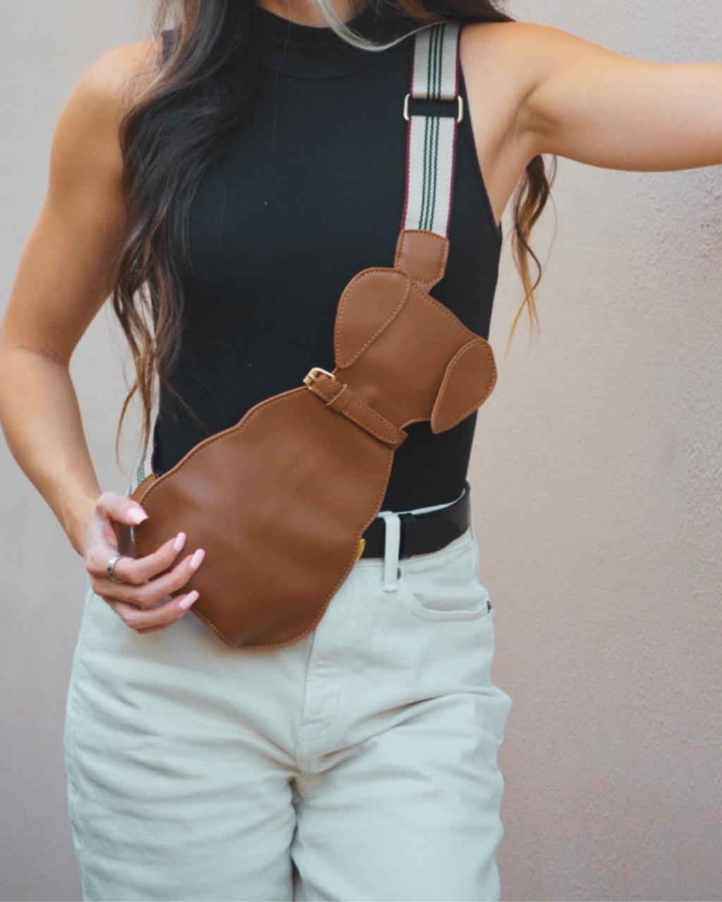 A woman wearing a brown dachshund-shaped shoulder bag with a white and black striped strap, paired with a black tank top and light beige pants.